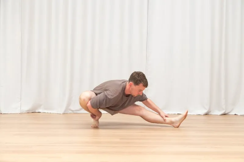 Keegan in a deep squat on one bent leg with the other leg stretched straight out to the side along the floor, reaching forward towards the extended foot