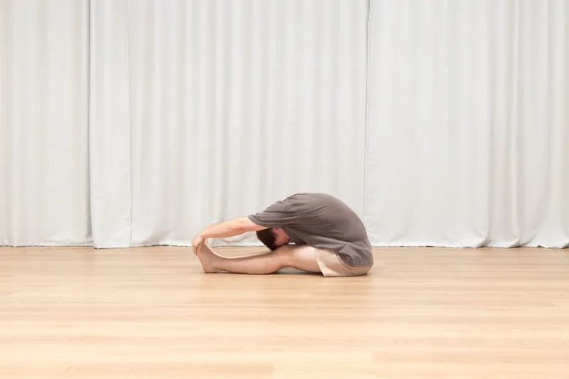 Keegan seated on a studio floor with his legs straight out in front of him, folded completely flat along them, chest and head resting on his thighs and both hands holding his feet