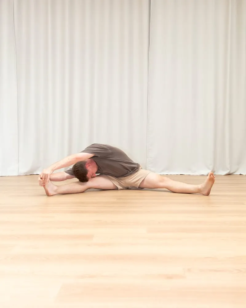 Keegan sitting with his legs split wide apart on the floor, folded sideways along one straight leg with both hands reaching to that foot