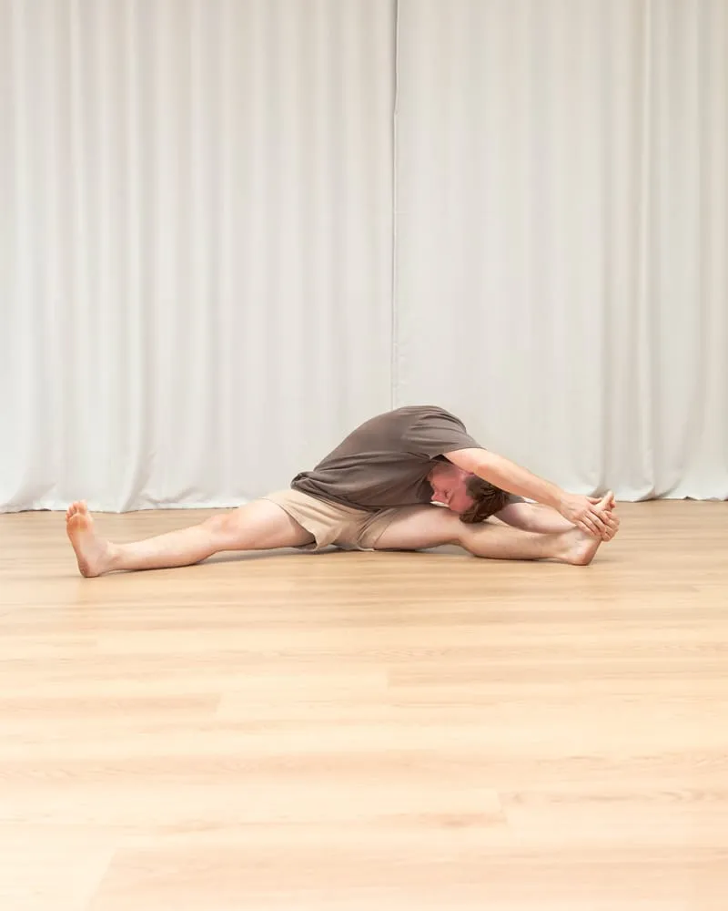 Keegan sitting with his legs split wide on the floor, folded along one leg with his chest low and both arms extended to the foot