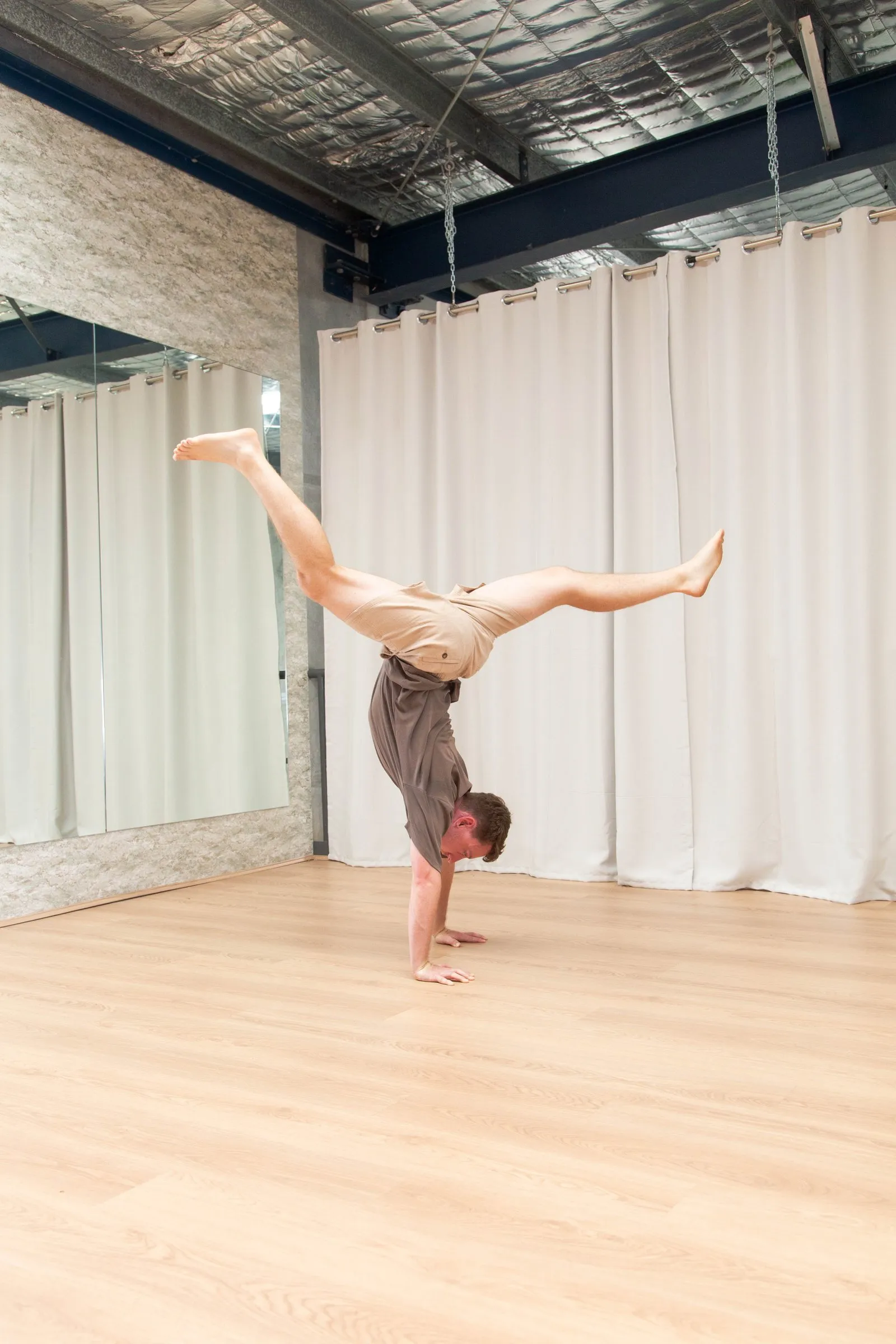 Keegan in a straddle handstand in a mirrored studio, weight on both hands with his legs split wide apart above him