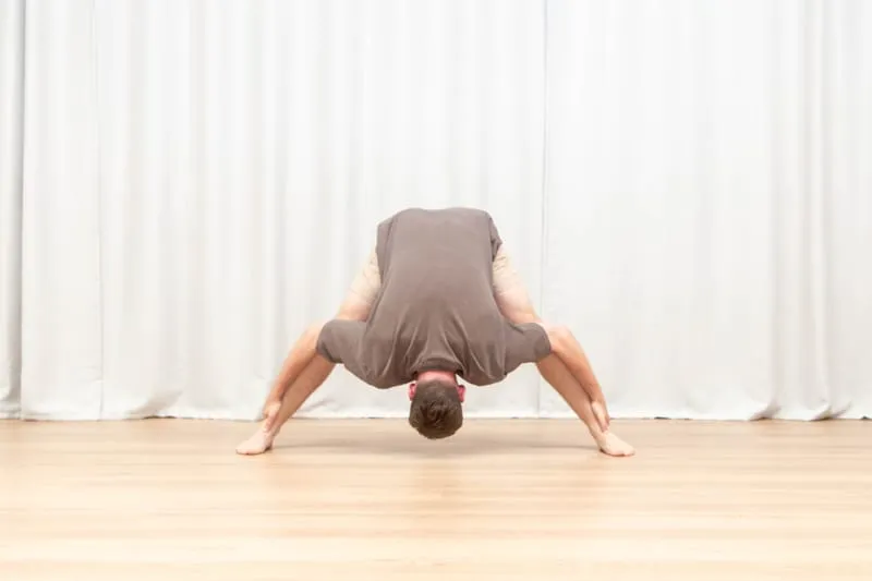 Keegan standing with his feet set wide apart, folded all the way forward with both palms flat on the floor and his head hanging between his legs