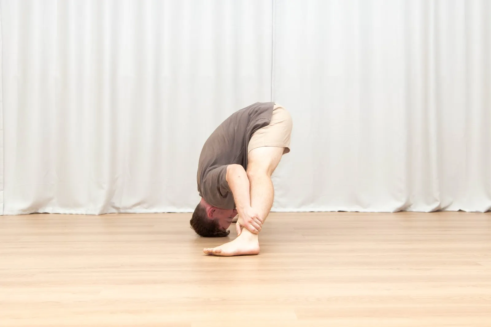 Keegan in a deep standing forward fold with straight legs, his head lowered all the way to the floor beside his foot and his hands clasped around his shin