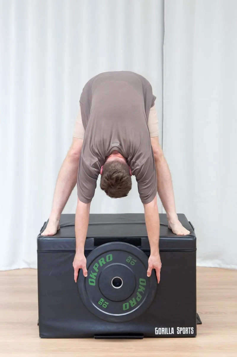 Keegan standing with his feet wide apart, folded forward over a padded gym box with both hands resting on a weight plate on top of it