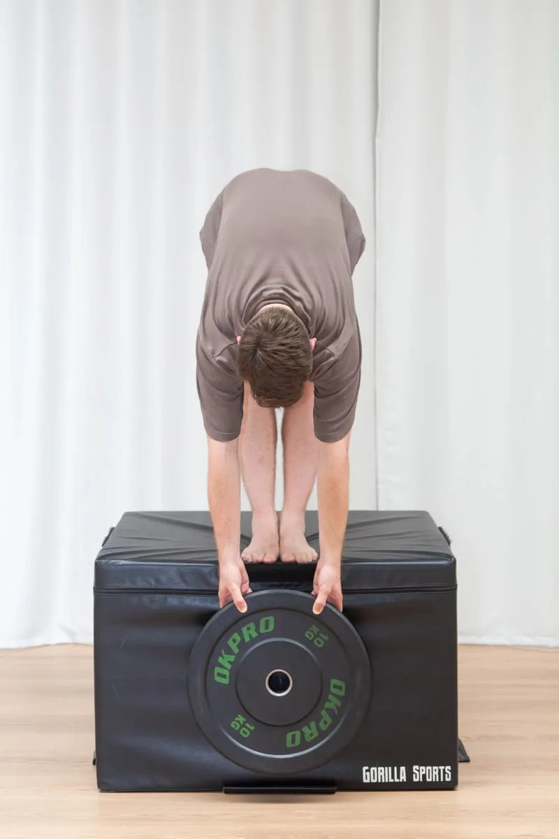 Keegan standing with his feet together, folded forward over a padded gym box with both hands on a weight plate resting on it