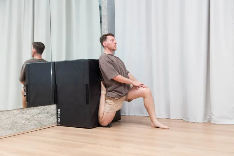 Keegan kneeling in a lunge with his back foot raised against a padded box behind him, torso upright, opening the front of the back hip