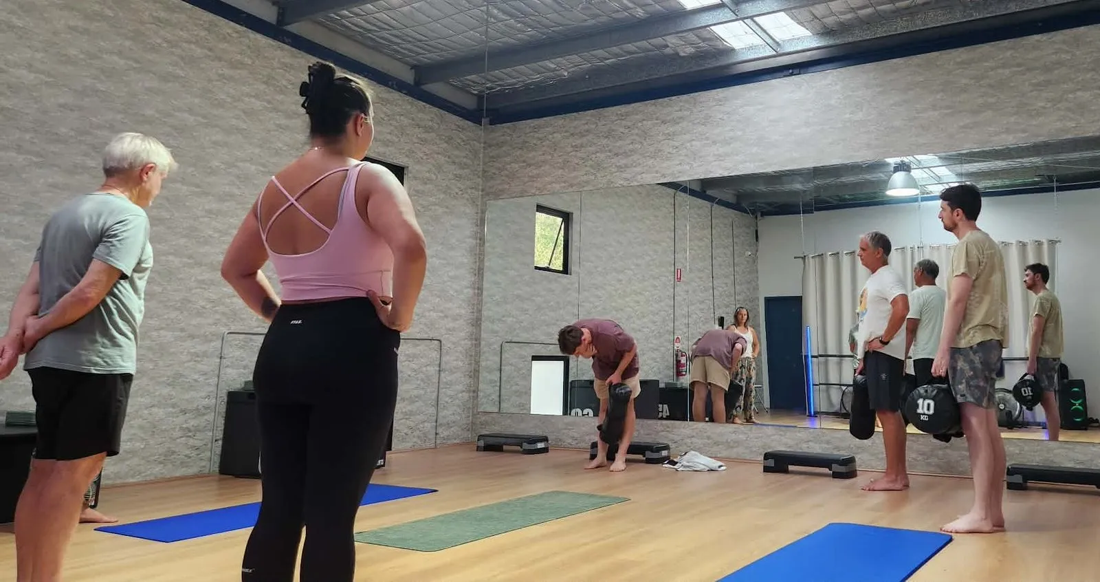 A group class in progress in a gym studio, participants working through a movement drill on mats while Keegan demonstrates