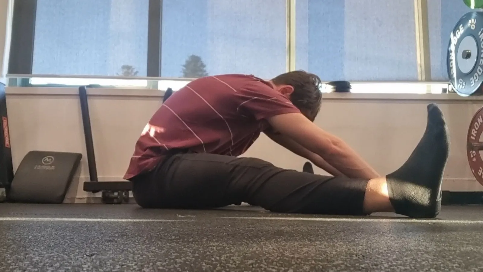 Keegan sitting on a gym floor with his legs straight out in front of him, folding forward, his chest still well clear of his thighs
