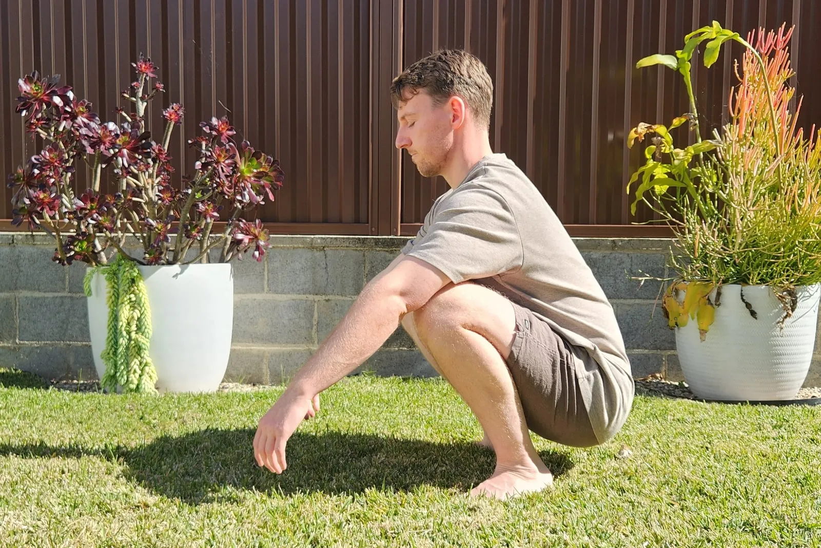 Keegan at the bottom of a deep squat on grass, seen from the side, with his hips below his knees and both heels flat on the ground.