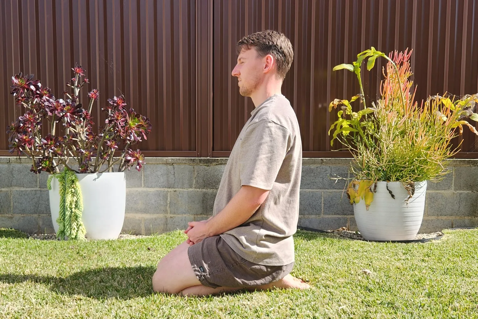 Keegan kneeling and sitting back onto his heels on grass, seen from the side, torso upright and the tops of his feet flat on the ground.