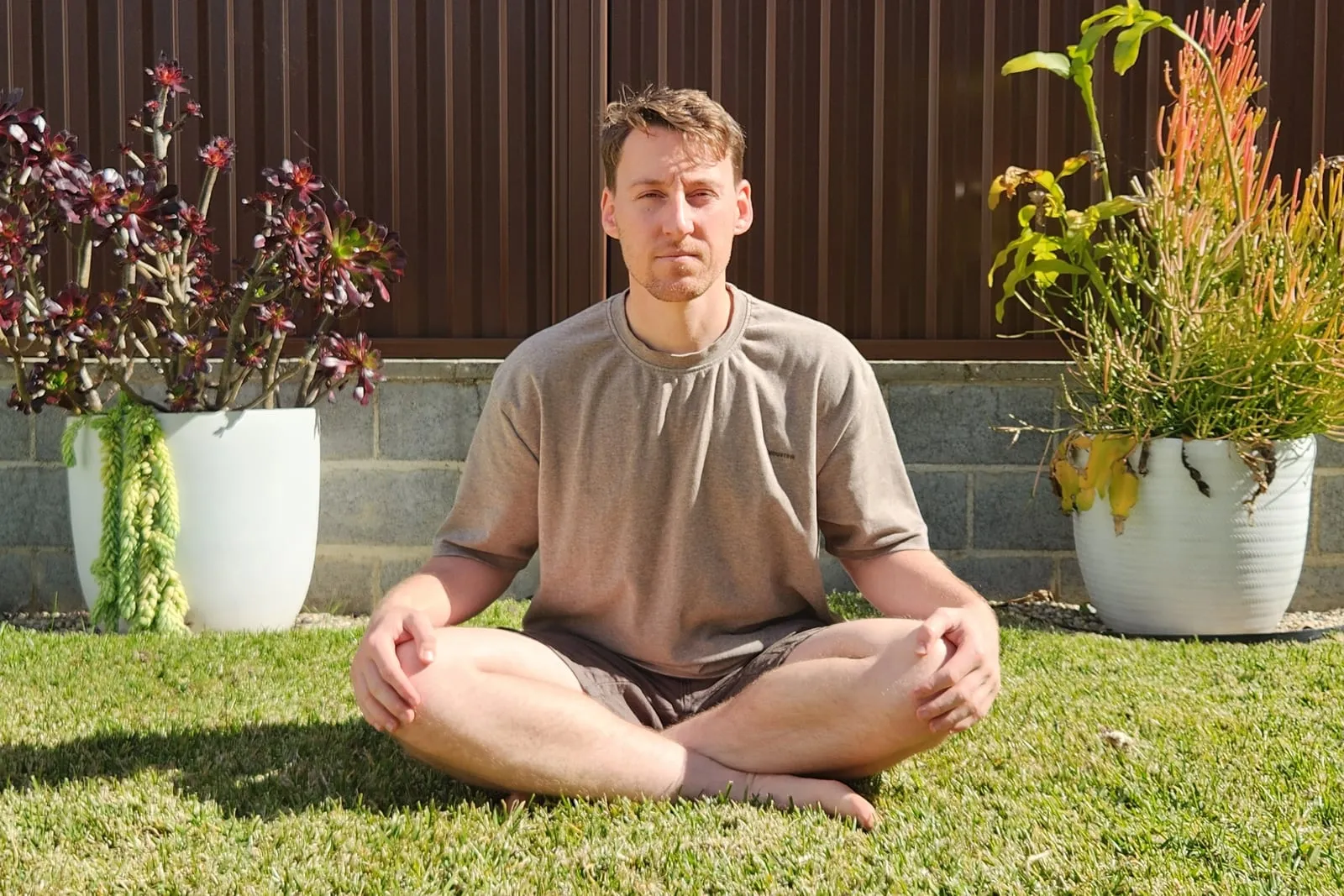 Keegan sitting cross-legged on grass, seen from the front, sitting tall with each foot tucked under the opposite leg and both knees resting low.