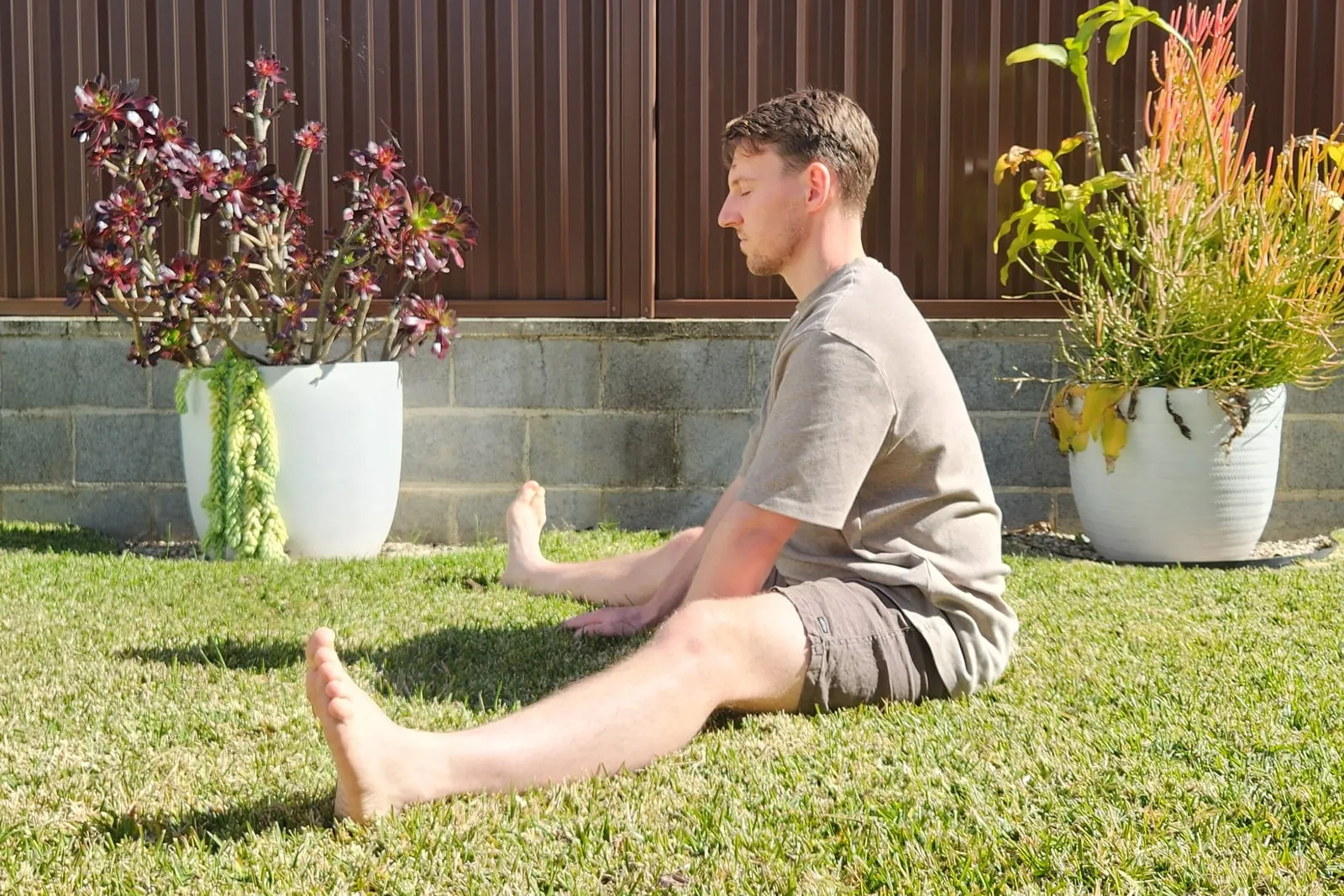 Keegan sitting in a wide straddle on grass, seen from the side, sitting tall with his pelvis upright rather than rolling backward.