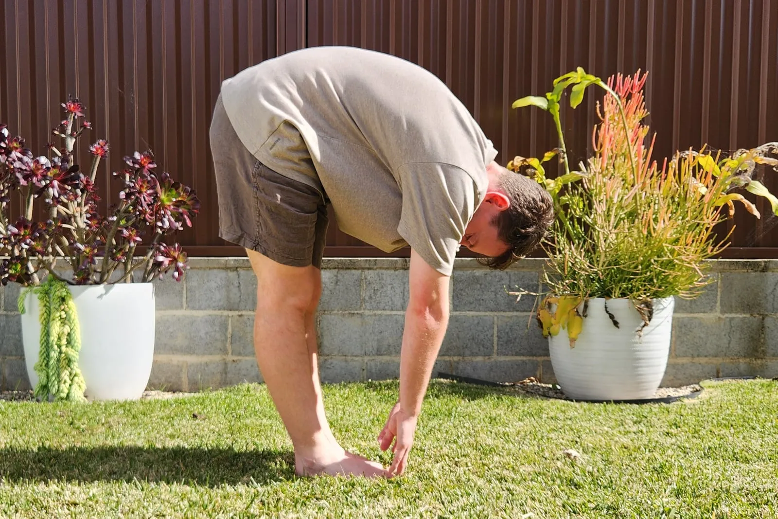Keegan bending forward from standing on grass, seen from the side, legs straight and his fingertips reaching down to the ground beside his feet.