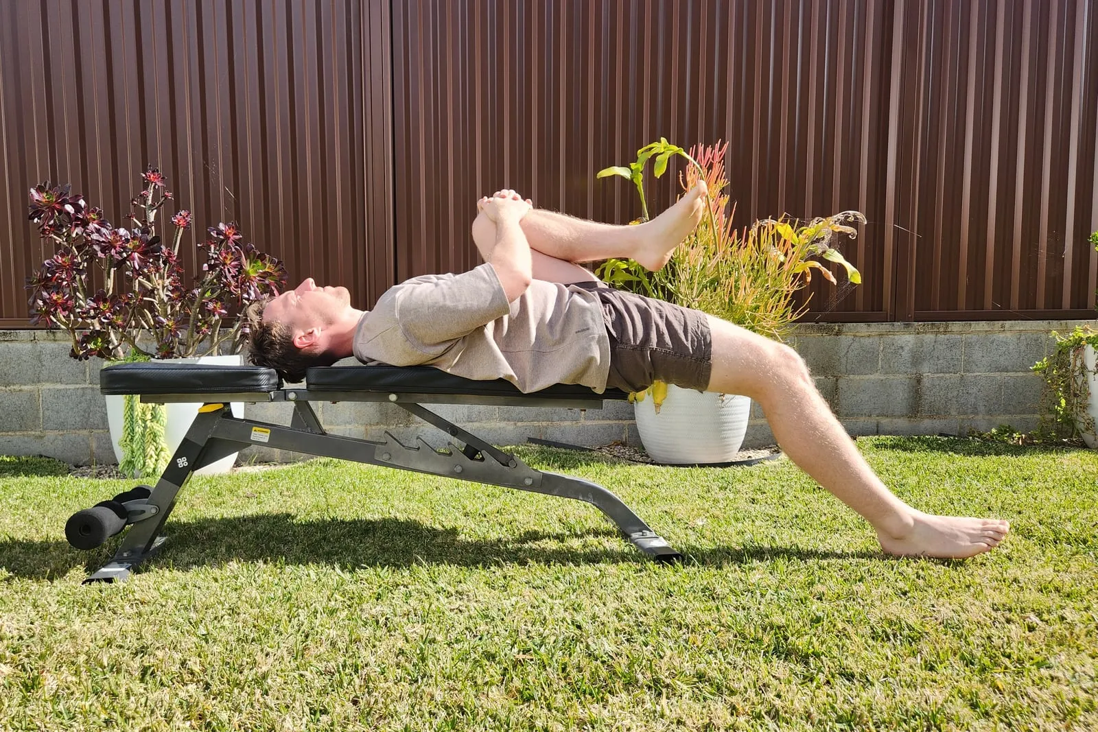 Keegan lying on his back at the end of a bench, one knee hugged to his chest and the other leg lowered so that thigh rests below the line of the bench.
